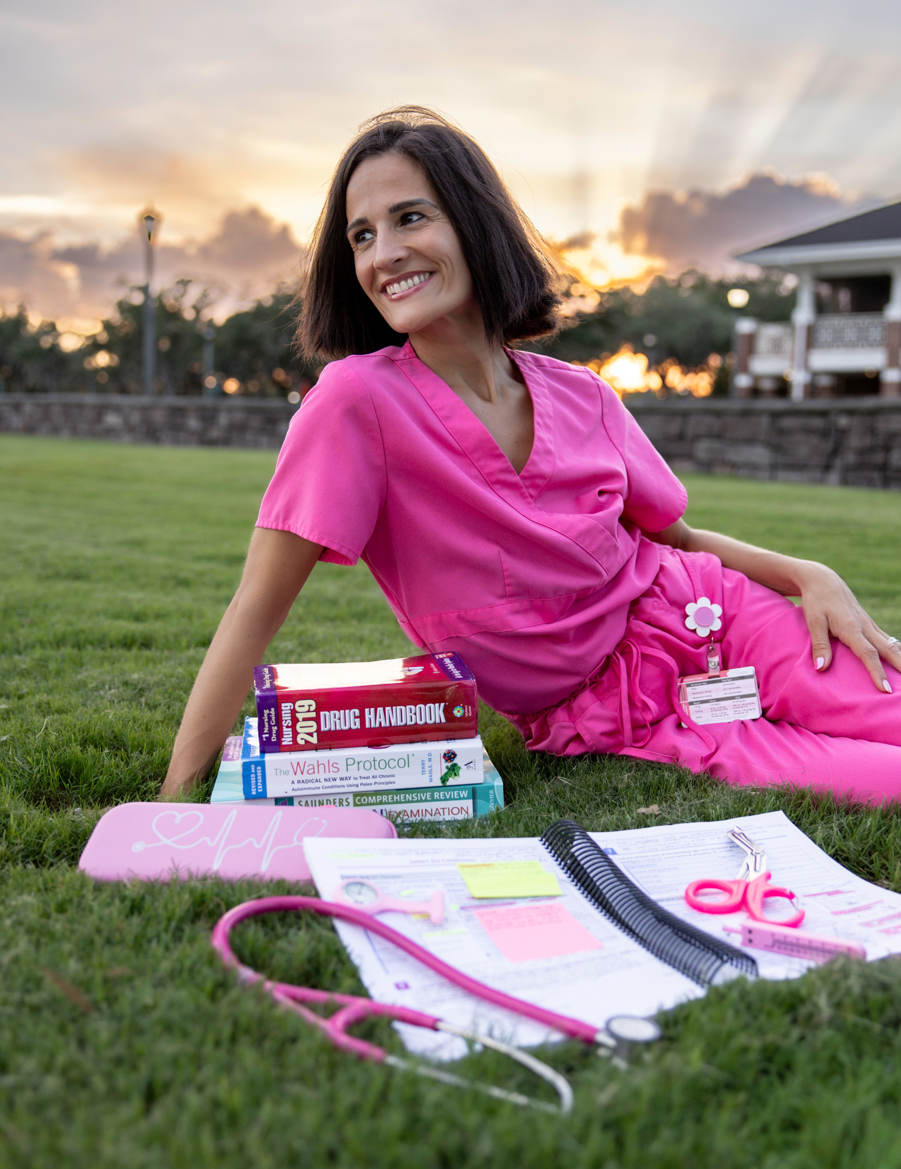 Camille Engle, FNP-C, sitting on grass in pink scrubs surrounded by nursing textbooks and study materials at golden hour